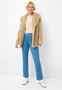 Manteau en beige duveteux avec un large col, porté sur un pull léger et un jean bleu, associé à des baskets blanches sur un fond blanc.