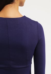 Mörklila, figursydd topp med rund halsringning och långa ärmar. Slät, texturerad tyg med subtila sömmar längs ryggen.