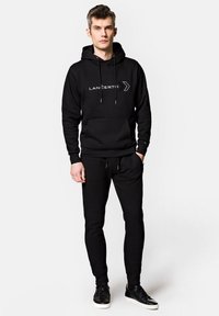Czarna hoodie z sznurkami i przednią kieszenią, z białym logo "LANGERTO". Połączona z dopasowanymi czarnymi dresami i czarnymi sneakersami.