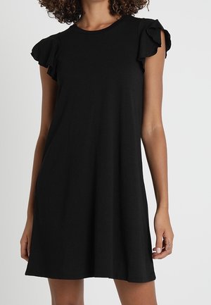 Femme portant une courte robe noire avec des manches courtes volantées, debout devant un fond clair uni.