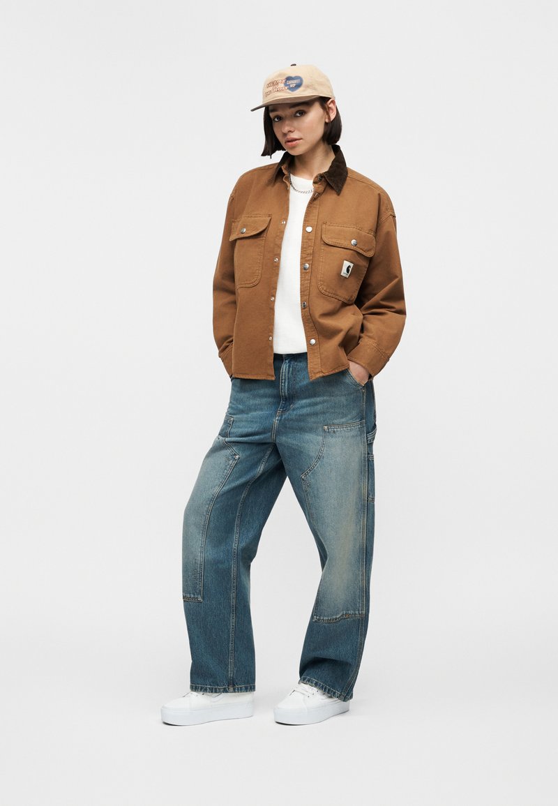 Giovane donna con un berretto beige, giacca marrone con bottoni, camicia bianca, jeans blu larghi e scarpe da ginnastica bianche, in piedi con le mani nelle tasche.