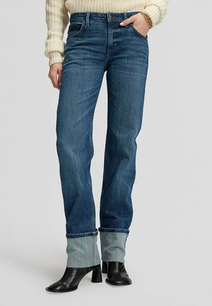 Femme portant un jean bleu retroussé, des bottines noires à talons, un pull en maille crème, avec une main dans la poche et un bracelet en or au poignet.