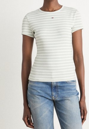 Osoba ubrana w jasnoszarą i białą koszulkę z krótkim rękawem w poziome paski oraz niebieskie jeansy z denimu, stojąca na jednolitym tle.