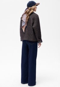 Brązowa kurtka o fakturze z szerokimi rękawami, zestawiona z ciemnoniebieskimi spodniami typu wide-leg. Wzorzysty szalik związany z tyłu oraz granatowa czapka.