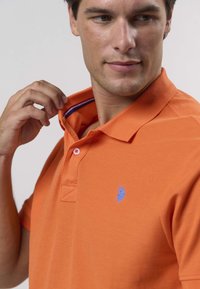 Oranž polo-särk tekstuuritud kangast, vasakul rinnal sinise logoga ja krae sees kontrastsete siniste detailidega. Kolme nupuga nööbiliist.