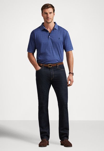 Άντρας όρθιος με τα χέρια στις τσέπες, φοράει μπλε πόλο με κοντά μανίκια, σκούρο τζιν, καφέ ζώνη, ρολόι και καφέ μοκασίνια, μπροστά σε απλό φόντο.