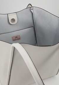 Valkoinen nahkatote-bag, jossa on sileä pinta, musta reuna ja beige sisustus, jossa on pieni tasku, jonka päällä on teksti "ANNA FIELD."