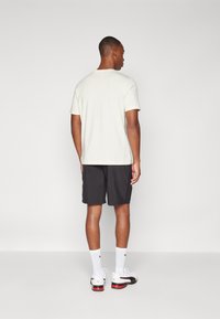 Camiseta de color claro con mangas cortas, combinada con pantalones cortos oscuros. Modelo vistiendo calcetines blancos y zapatillas negras y rojas, visto desde atrás.