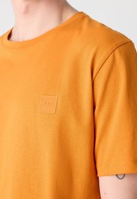 Personne portant un t-shirt orange uni avec un petit logo en relief portant l’inscription "BOSS" sur le côté gauche de la poitrine et un tatouage partiel visible sur le bras.