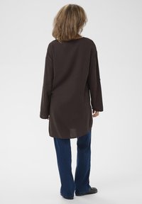 Camicia a maniche lunghe marrone scuro con vestibilità rilassata, caratterizzata da un collo arrotondato e spacchi sui lati, abbinata a jeans a gamba larga blu.