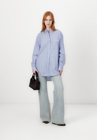 Modra in bela prugasta srajca z ovratnikom na gumb, dolgimi rokavi in žepom na prsih, v kombinaciji s svetlimi zvonastimi jeans hlačami in črno torbico.