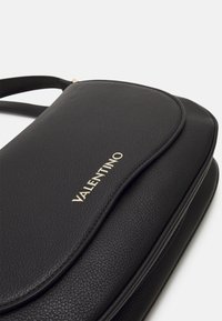 Sac à main en cuir noir avec une texture lisse, présentant un logo doré et un design de rabat courbé. Sangle simple pour un port croisé ou à l'épaule.