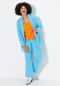 Un costume bleu clair comprenant une veste cintrée et un pantalon évasé, assorti d'un gilet orange et de bottines argentées. Tissu texturé avec des accents brillants.