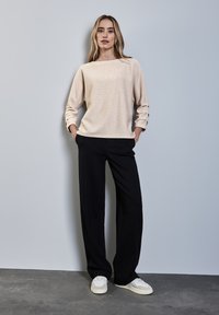 Maglione beige chiaro testurizzato con scollatura a barca, abbinato a pantaloni neri larghi e sneakers bianche, in piedi contro uno sfondo grigio.