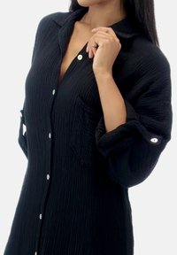 Camisa negra abotonada de tela arrugada con cuello, un bolsillo en el pecho y mangas remangadas con detalle de botón.