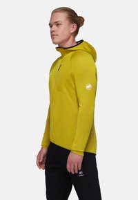 Gul huva jacka med texturerat tyg, zip-fickor på bröstet och kontrasterande svarta detaljer. Bärs med svarta byxor med en logo.