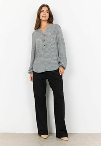 Blouse à motifs noir et blanc avec des manches longues et un patte de boutonnage, associée à un pantalon noir ample et des chaussures beiges à talon pointu.