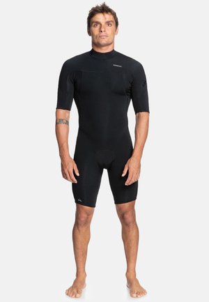 Man staat blootsvoets, draagt een zwarte korte mouw, knielange wetsuit met een tatoeage op zijn linker onderarm.