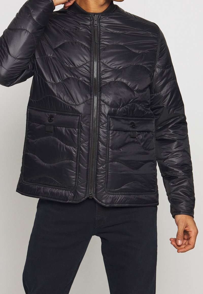 Giubbotto impermeabile nero con texture trapuntata, zip frontale e due tasche sul petto con chiusura a bottone. Design leggero e vestibilità regolare.