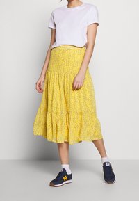 Person iført en hvid kortærmet top, gul blomstret, lagdelt midiskørt, hvide sokker og mørkeblå New Balance sneakers.