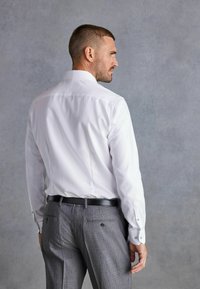 Weißes Hemd mit Knöpfen, taillierte Passform, glatte Textur, kombiniert mit grauen gemusterten Hosen, ergänzt durch einen schwarzen Gürtel und dezente Taschendetails.