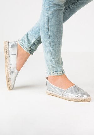 Personne portant un jean bleu clair et des espadrilles plates argentées à paillettes avec des semelles en jute sur un fond blanc.