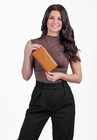 Tan leather wallet with a sleek, elongated design. Features a small logo and a smooth texture, held by a person wearing a form-fitting top.