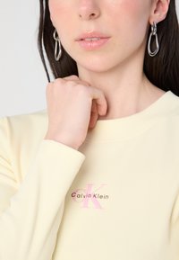 Žuta rebrasta majica dugih rukava s roza Calvin Klein logotipom. Model nosi srebrne apstraktne naušnice, a na njenoj koži je vidljiva glatkoća i prirodan izgled šminke.