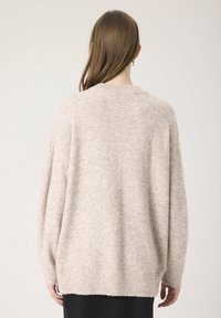 Femme avec de longs cheveux bruns portant un pull en laine beige ample et une jupe noire, vue de dos sur un fond uni.