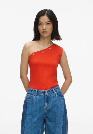 Femme portant un haut rouge asymétrique avec des œillets métalliques et un jean bleu ample, debout les mains derrière le dos.