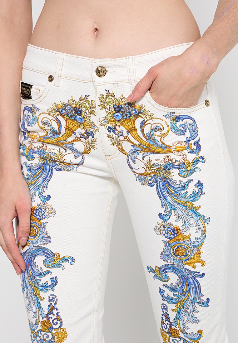 Weiße Jeans aus Denim mit aufwendiger blauer und goldener Blumenstickerei an den Beinen, akzentuiert mit goldenen Hardware-Elementen und Taschen.