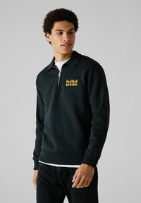 Zwarte rits-hoodie gemaakt van zachte stof, met een hoge kraag en een zichtbaar gele "Red Bull Racing" logo op de borst.