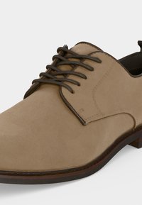 Chaussure habillée pour homme en daim beige avec garniture en cuir marron, lacets marron foncé et petit talon sur fond blanc.