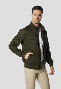 Kurtka oliwkowa z lekkiego materiału, z zamkiem błyskawicznym z przodu, dwoma kieszeniami na piersi oraz ściągaczami na rękawach i na dole.