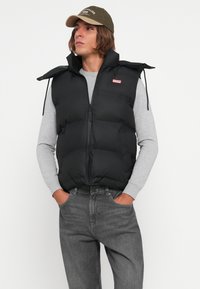Melnā pūkainā veste ar augstu apkakli un rāvējslēdzēja aizdari. Valkāta virs pelēka garām piedurknēm krekla. Redzamās džinsu bikses pie jostas.
