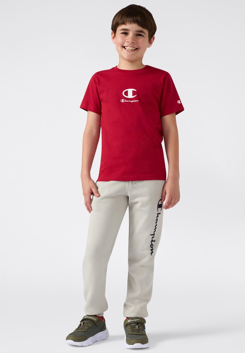 Junge lächelt, trägt ein rotes Champion-T-Shirt, hellgraue Jogginghose mit Logo und olivgrüne Champion-Sneaker, steht vor weißem Hintergrund.