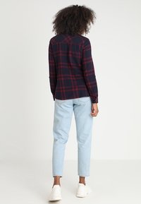 Marinegeruite flanellen shirt met rode accenten, lange mouwen en een relaxte pasvorm, gecombineerd met lichtblauwe spijkerbroek en witte sneakers.