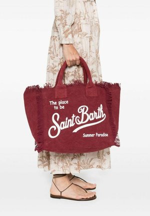 Persoon die een beige jurk met patroon en sandalen draagt, houdt een grote rode tas met franjes vast met de tekst "Saint Barth, Summer Paradise."