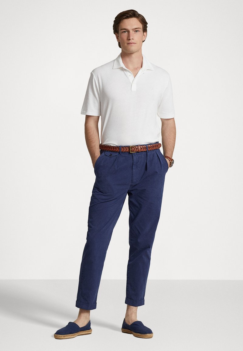 Witte polo, marineblauwe broek, een bruine gevlochten riem en marineblauwe espadrilles. Eenvoudig ontwerp, ontspannen pasvorm en opgerolde mouwen van de broek.