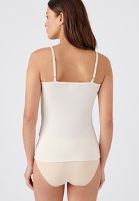 Canotta beige con spalline sottili, realizzata in morbido cotone, caratterizzata da un design aderente. Texture liscia, che si estende fino alla vita, senza decorazioni.