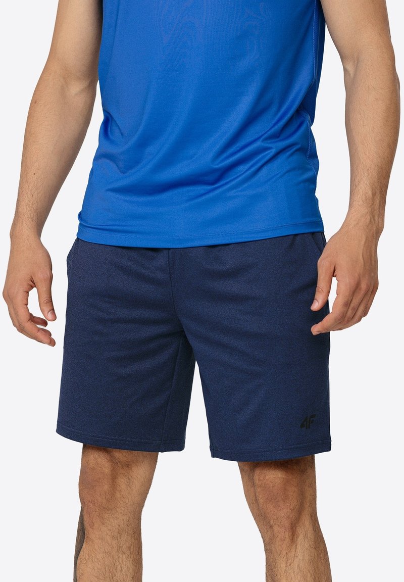 Homme portant un short de sport bleu et un débardeur bleu assorti, debout les bras détendus le long du corps.