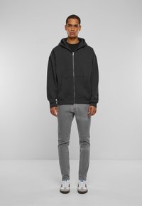 Felpa con zip grigia scura e cappuccio, con tasca a marsupio, abbinata a jeans slim-fit grigi chiari e sneakers bianche con dettagli blu.