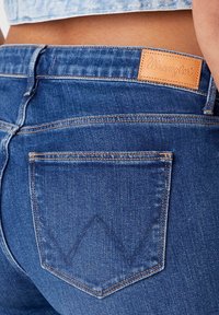 Detailní záběr zadní kapsy a pásku modrých džín Wrangler nošených s vykrojenou světle modrou košilí.