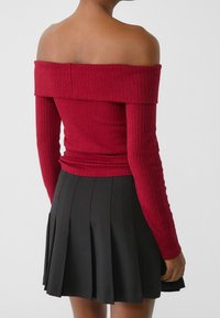 Haut rouge côtelé épaules dénudées à manches longues, associé à une jupe plissée noire. Le haut présente une coupe ajustée et un tissu texturé.