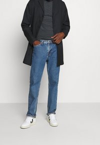Manteau long noir sur un pull en laine gris texturé, avec un jean en denim bleu et des baskets blanches avec des accents noirs.