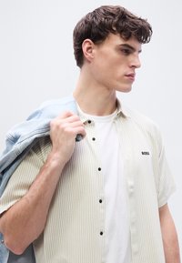 Νεαρός με κοντά καστανά σγουρά μαλλιά φορά ριγωτό κοντομάνικο πουκάμισο πάνω από λευκό μπλουζάκι, κρατώντας ανοιχτόχρωμο τζιν μπουφάν στον ώμο, κοιτάζει προς τα δεξιά.