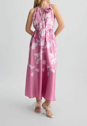 Vrouw draagt een roze mouwloze jurk met bloemenprint, een taillekoord en roosdetail, gecombineerd met hoge hakken sandalen versierd met veren.