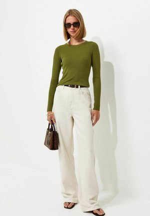 Mujer que lleva una camiseta verde de manga larga, pantalones anchos blancos, cinturón negro, gafas de sol, lleva un bolso marrón, de pie sobre un fondo blanco.