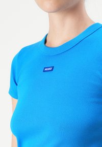 Close-up van een persoon die een felblauwe geribbelde ronde hals t-shirt draagt met een klein "HUGO" logo op de borst.