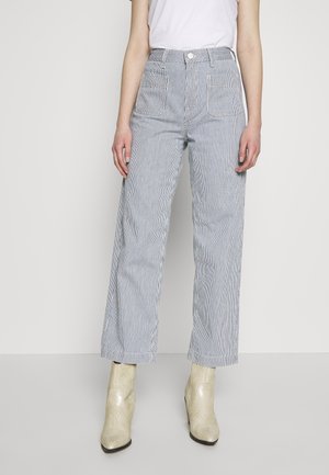 Femme portant un pantalon taille haute, à jambes larges, rayé bleu clair et blanc avec poches avant, un haut blanc sans manches rentré, et des bottines crème.